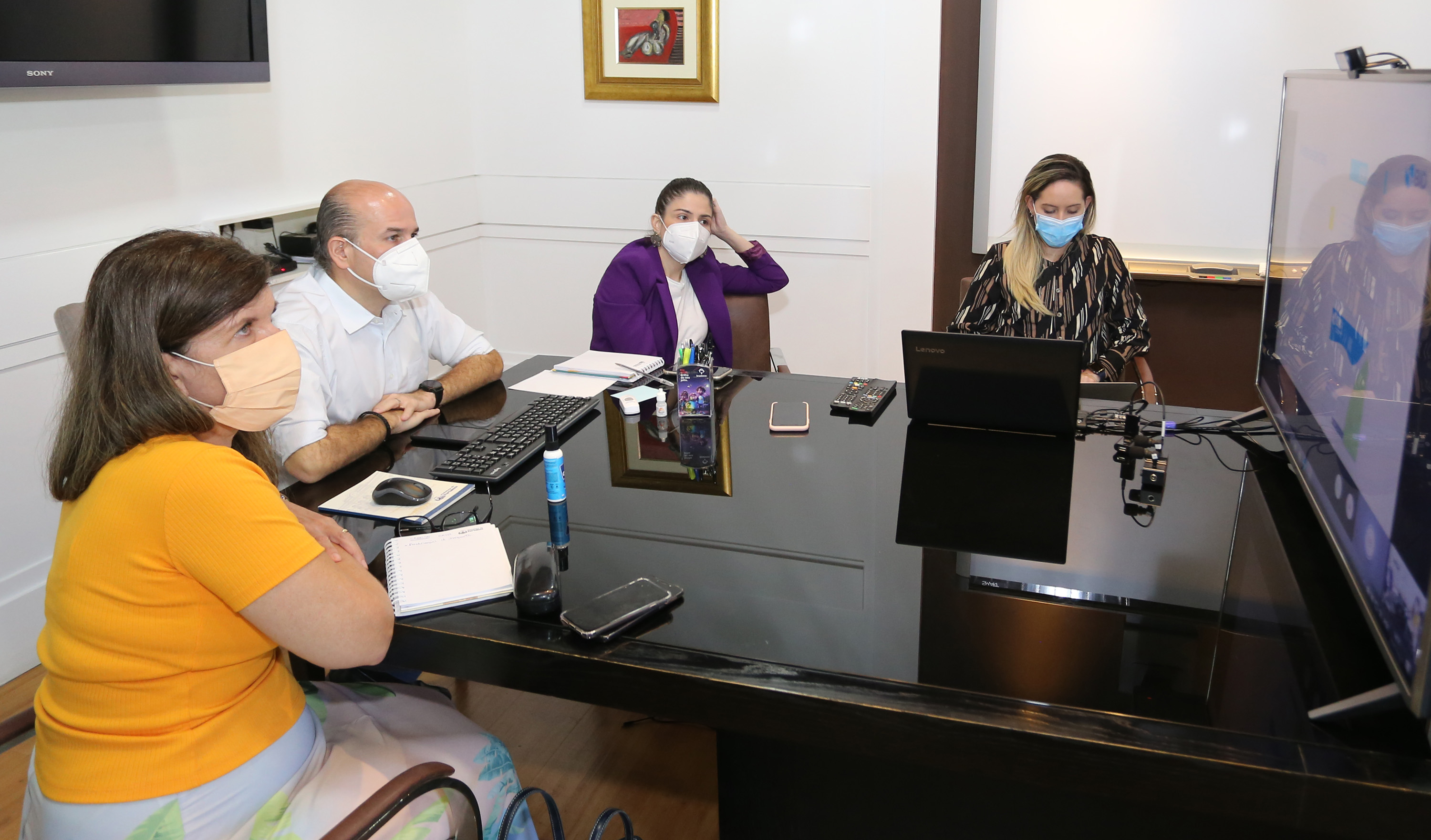 secretária Joana, prefeito, primeira-dama e moça loira sentados ao redor de mesa usando máscaras e olhando para monitor à frente